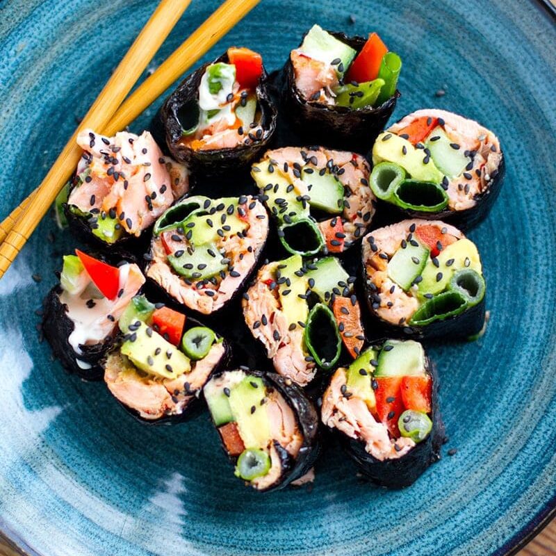 A colorful plate of keto sushi rolls wrapped in nori, garnished with avocado, sesame seeds, and spicy aioli.